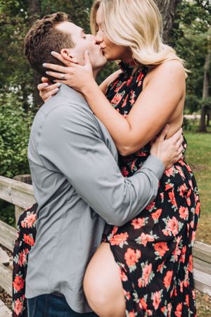 A romantic couple sharing a passionate kiss outdoors, with her wearing a floral dress and him in a gray shirt.