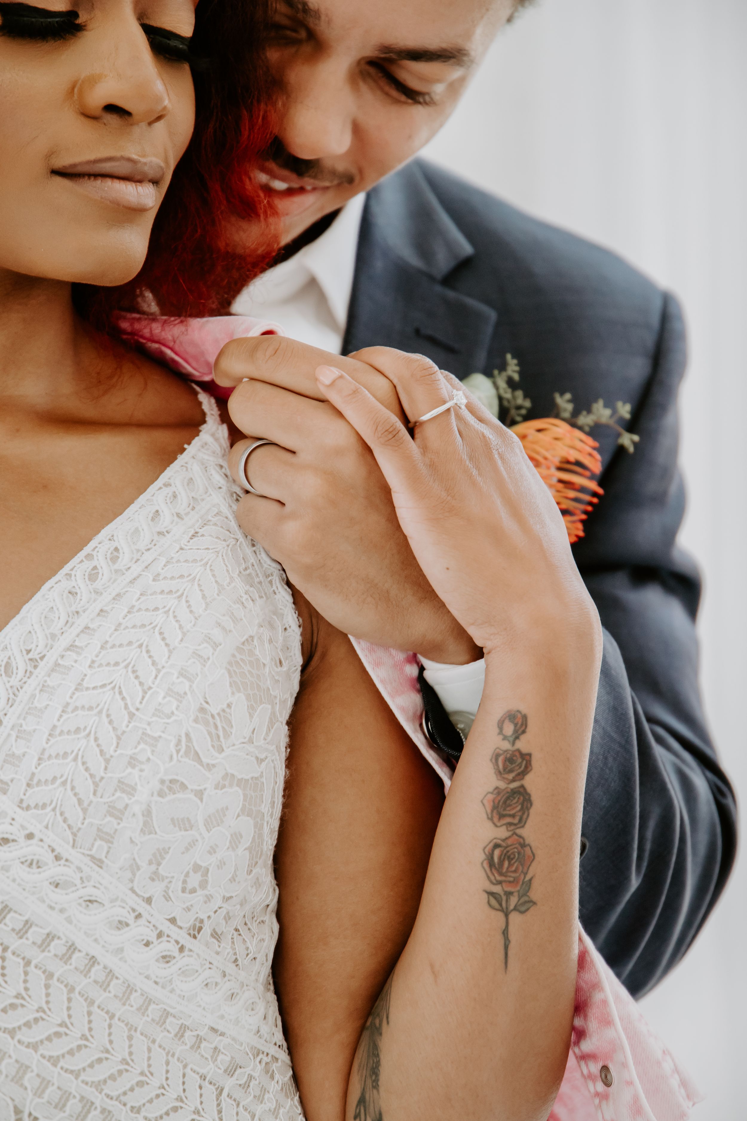 Detail shot of rose tattoo on arm with white lace wedding dress in background.