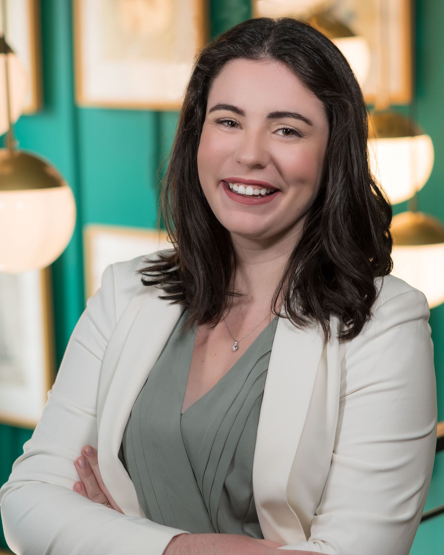 A professional headshot of a woman in a white blazer and grey top against a teal wall.