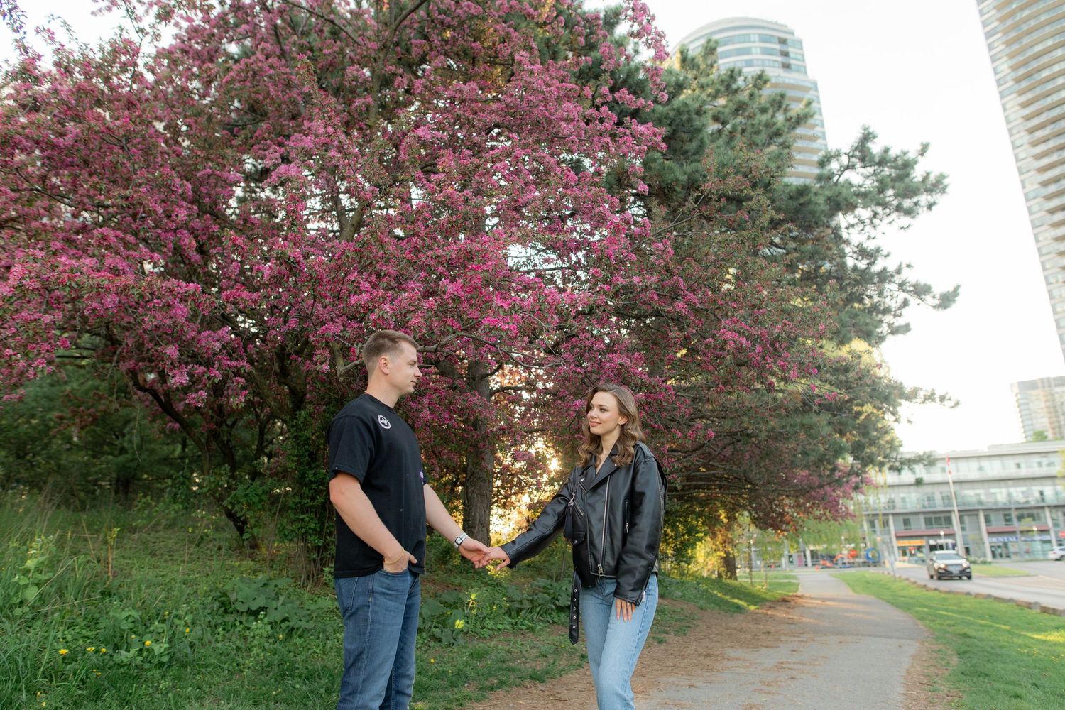 A couple holds hands beneath a flowering cherry blossom tree with urban buildings in the background.