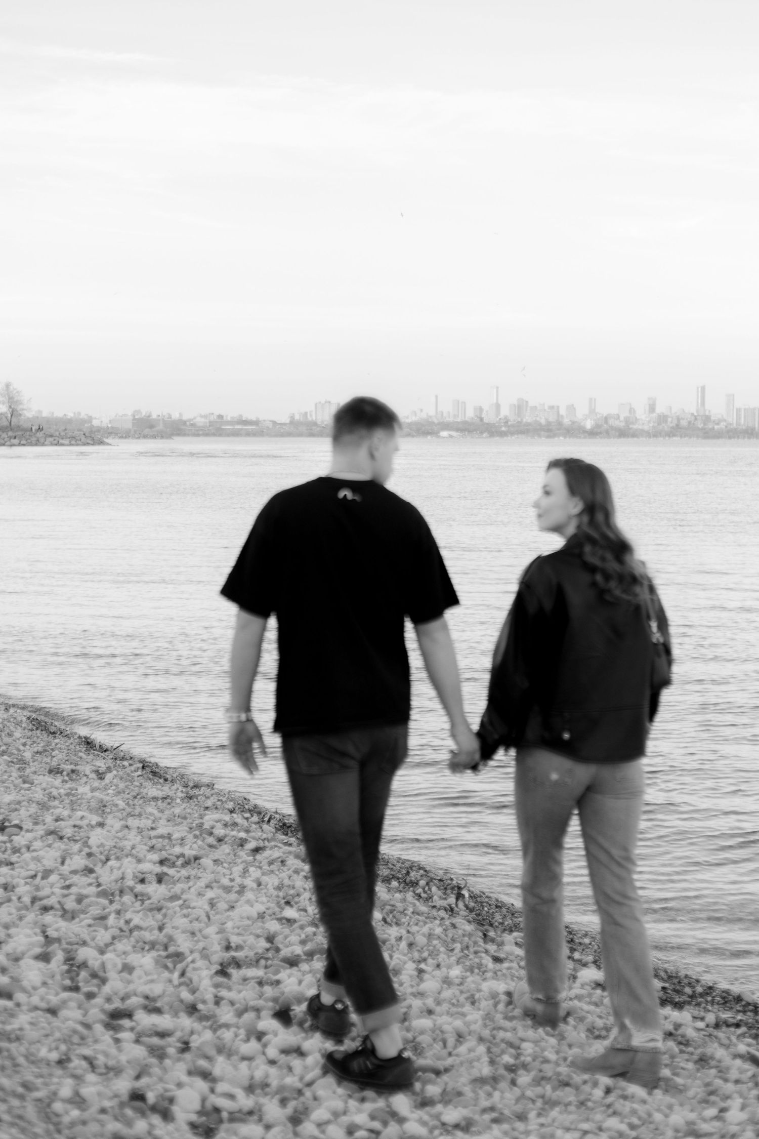 A couple walks along a rocky shoreline with a city skyline in the distance, captured in black and white.