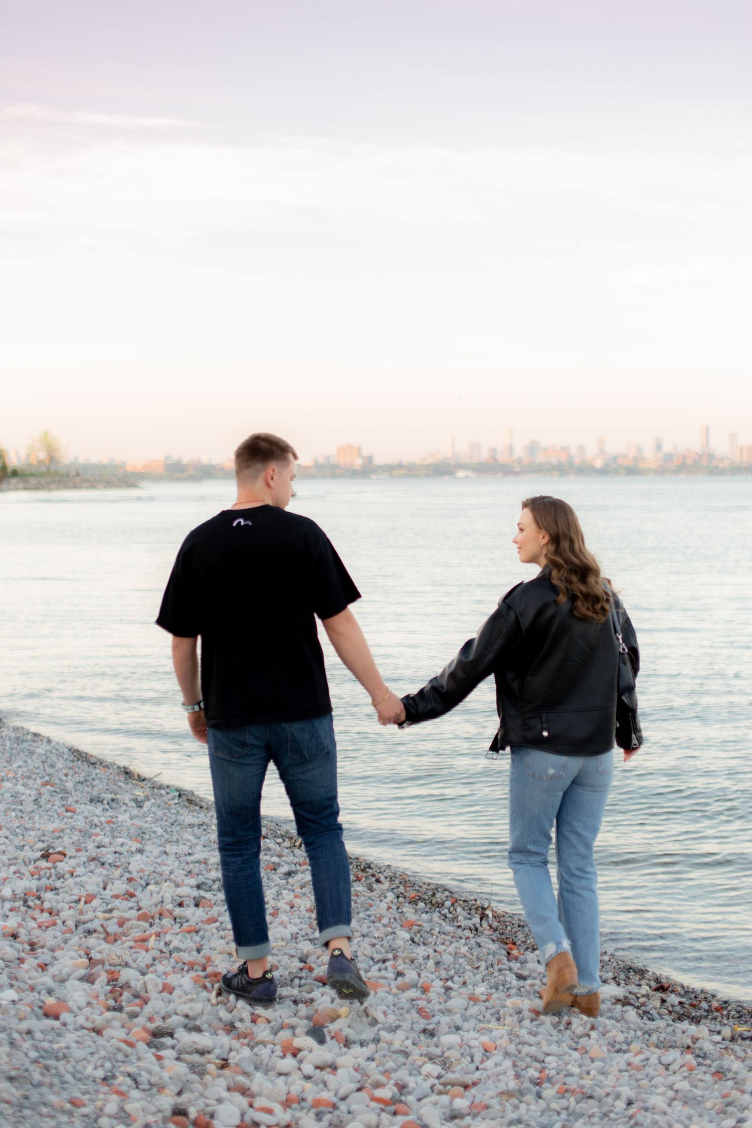 A couple strolling on a pebbly beach at sunset with a distant cityscape behind them.