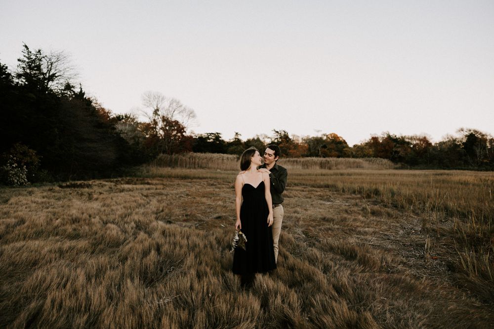 Couple embracing in a golden wheat field at dusk with colorful autumn trees in background. Miranda Photo & Co