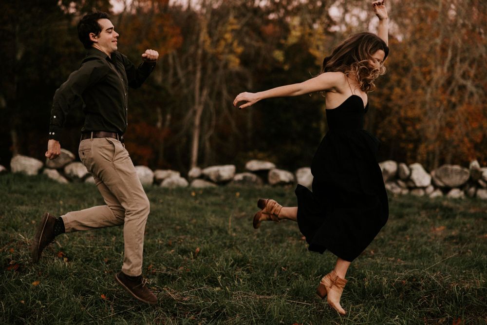 Playful couple running and dancing in an autumn garden with stone wall backdrop. Miranda Photo & Co
