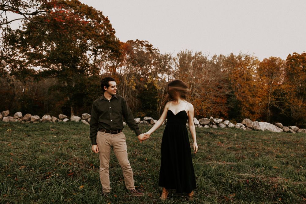 Couple holding hands in autumn field with colorful fall foliage, woman in black velvet dress, man in dark shirt. Miranda Photo & Co