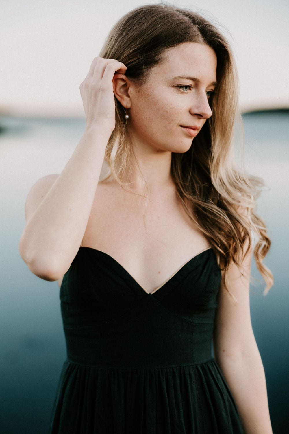 Beautiful woman in black strapless dress gently touching her hair near a tranquil lake. Miranda Photo & Co