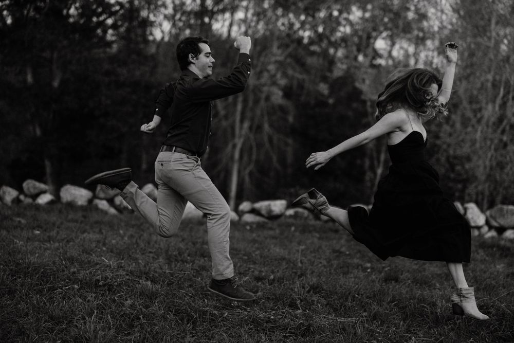 Black and white photo of a couple joyfully jumping and running together on a grassy lawn outdoors. Miranda Photo & Co