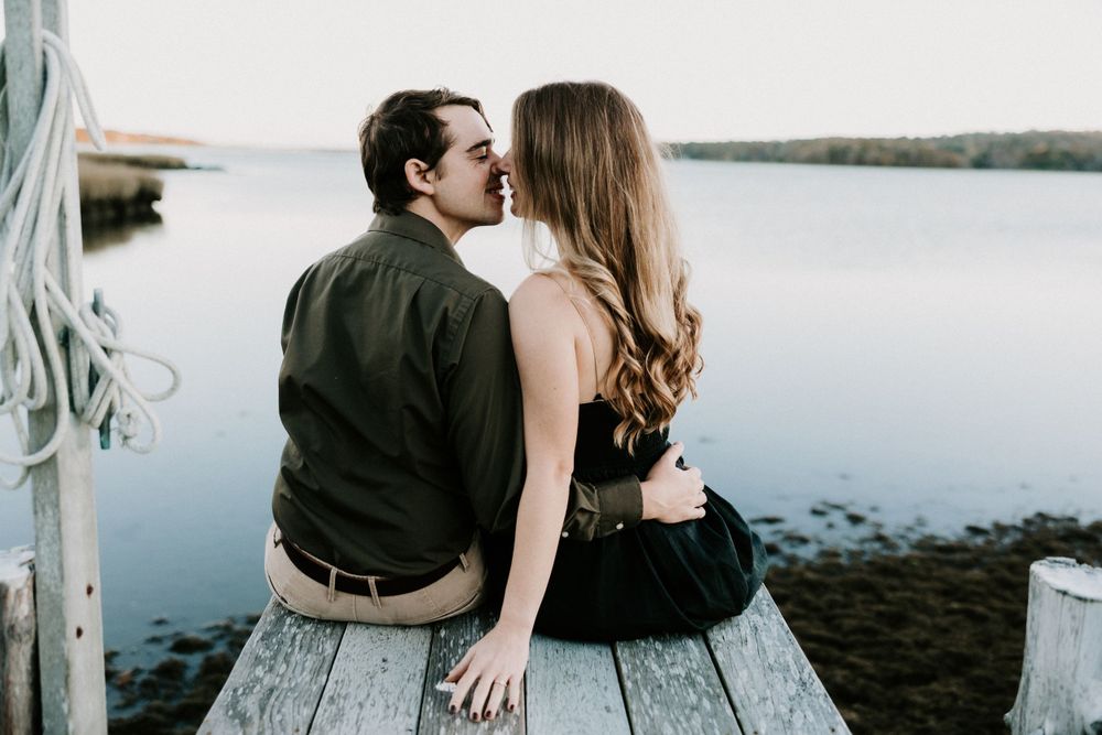 Couple sharing a kiss while sitting on a wooden dock overlooking a serene coastal waterway. Miranda Photo & Co