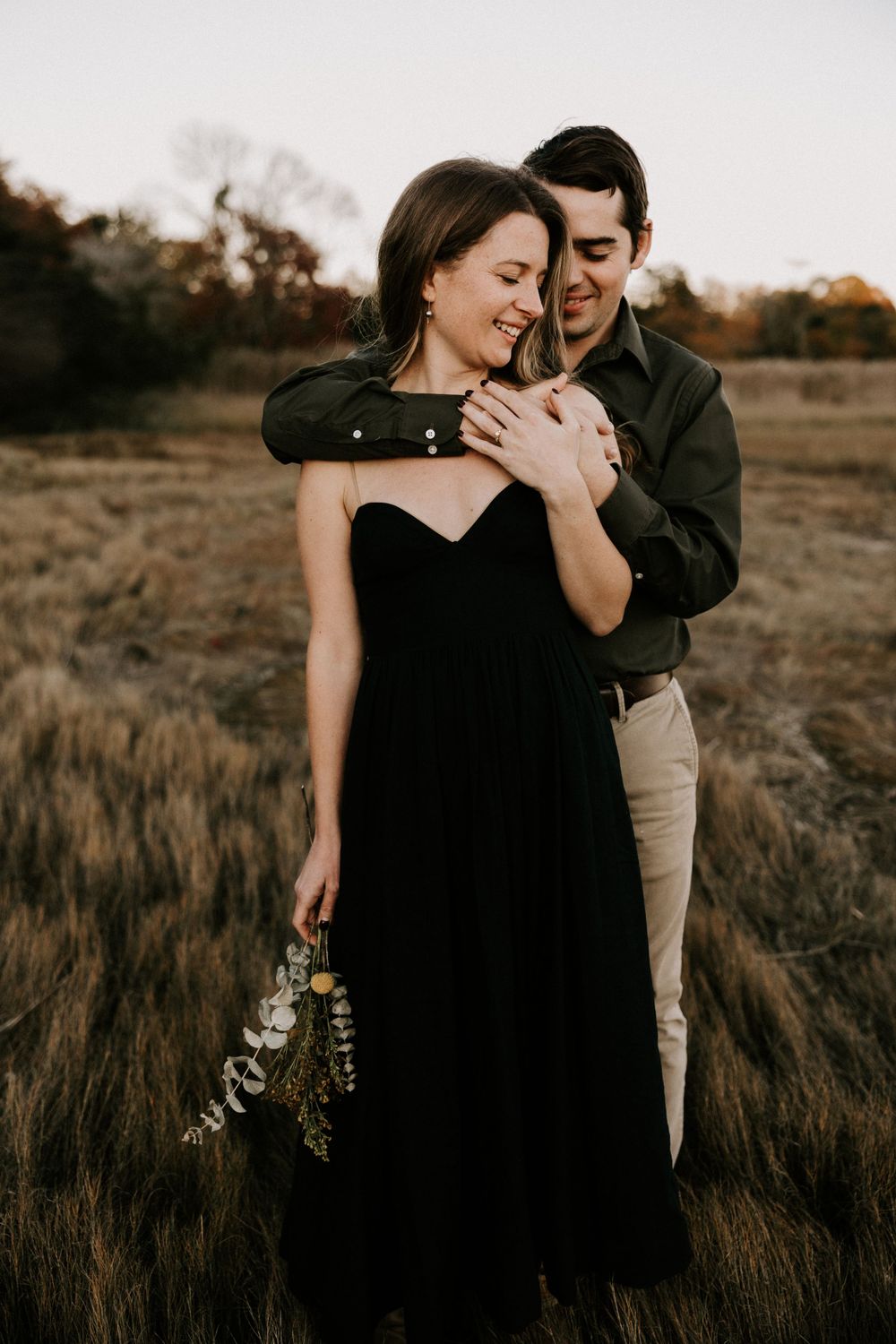 Man embracing woman from behind in golden field at sunset, woman smiling holding wildflowers. Miranda Photo & Co