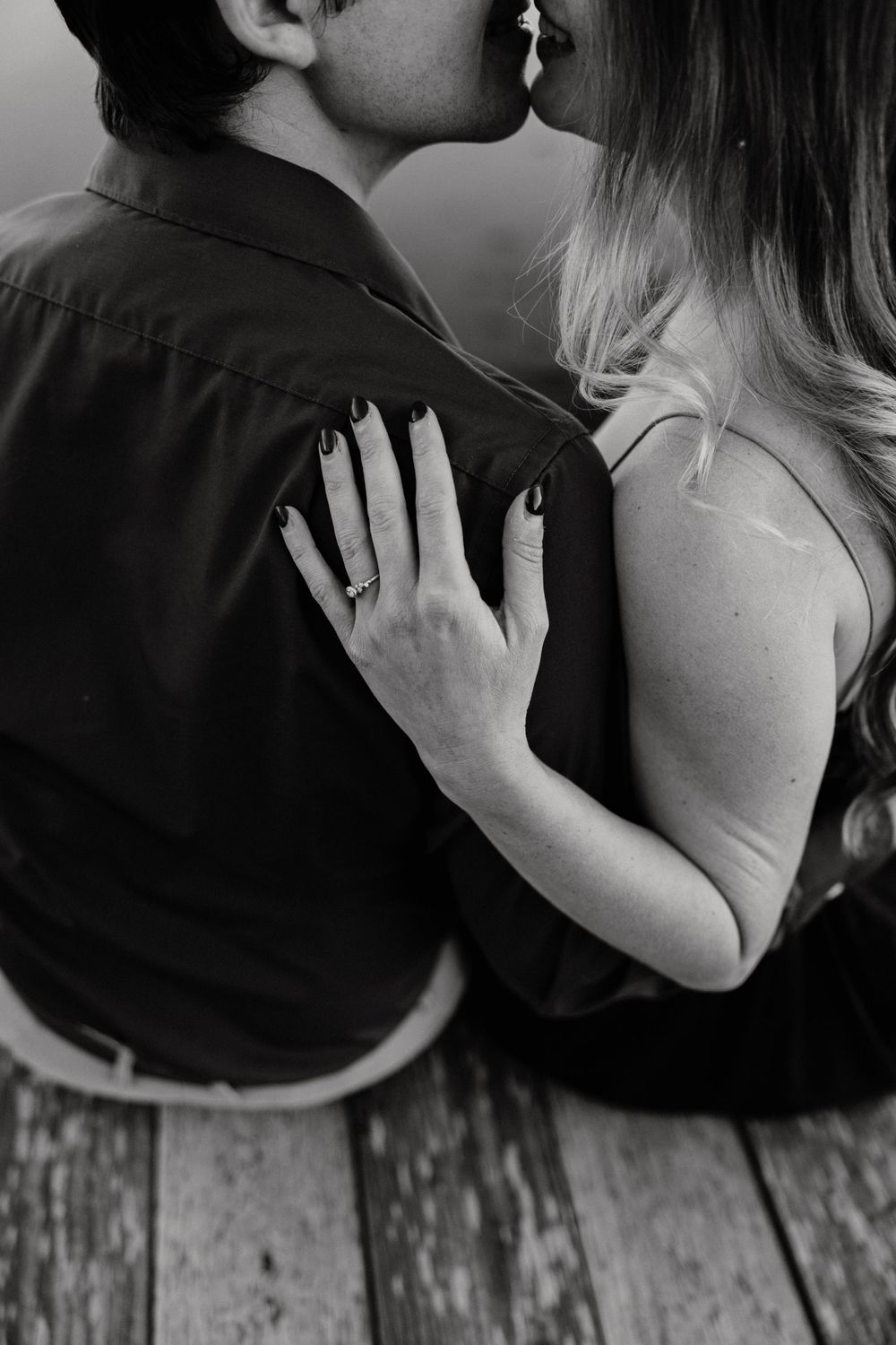 Black and white photo of a couple kissing, woman showing engagement ring on man's shoulder while sitting on wooden dock. Miranda Photo & Co