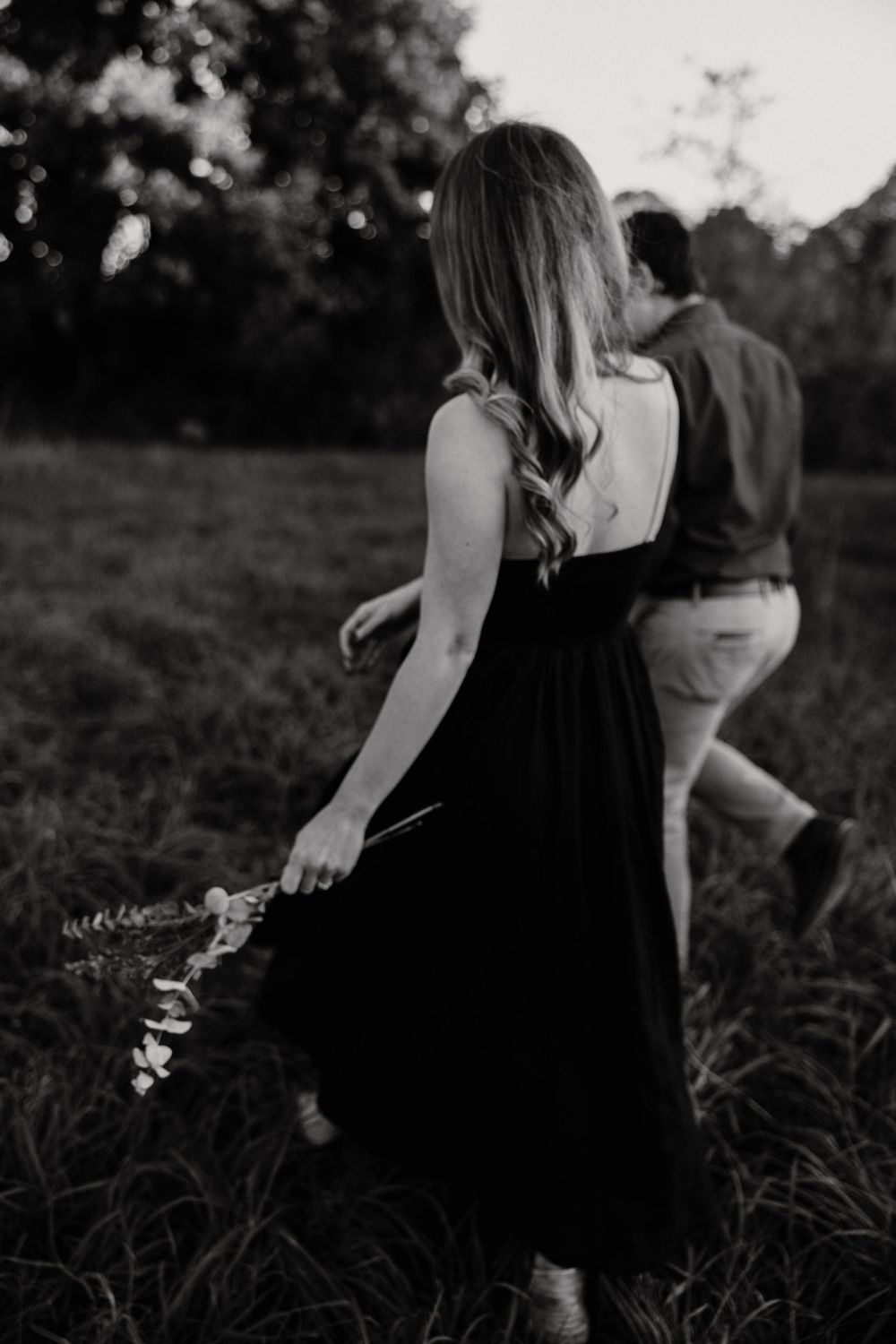 Black and white photo of couple walking through grassy field, woman in black dress holding flowers. Miranda Photo & Co