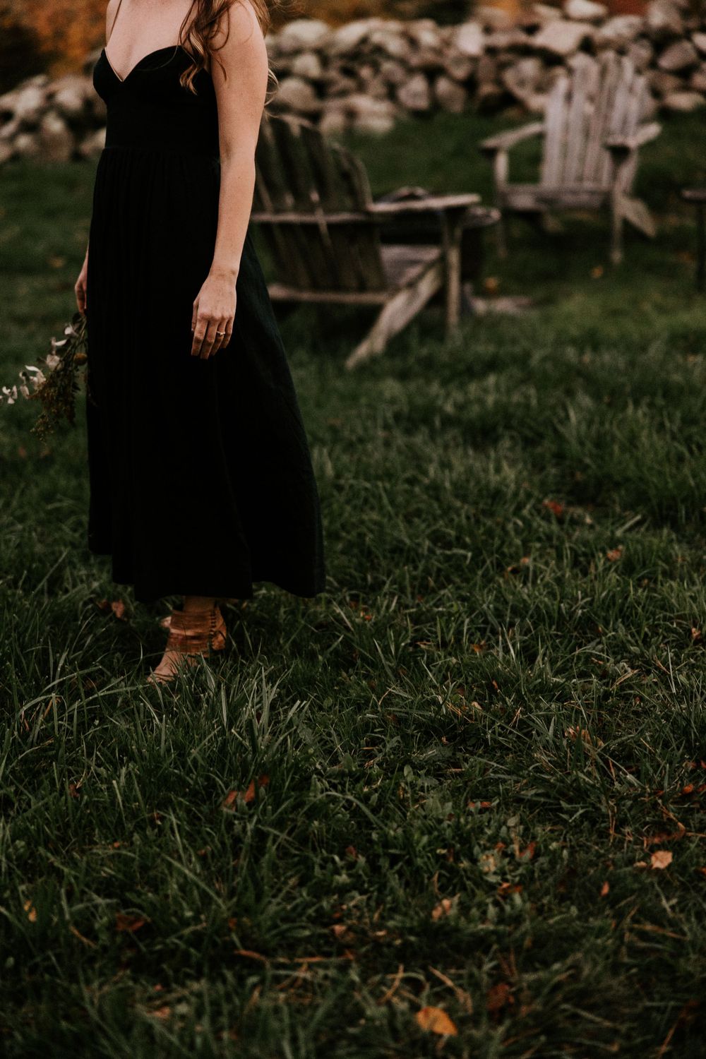 Woman in elegant black strapless dress standing on green grass near wooden Adirondack chairs. Miranda Photo & Co
