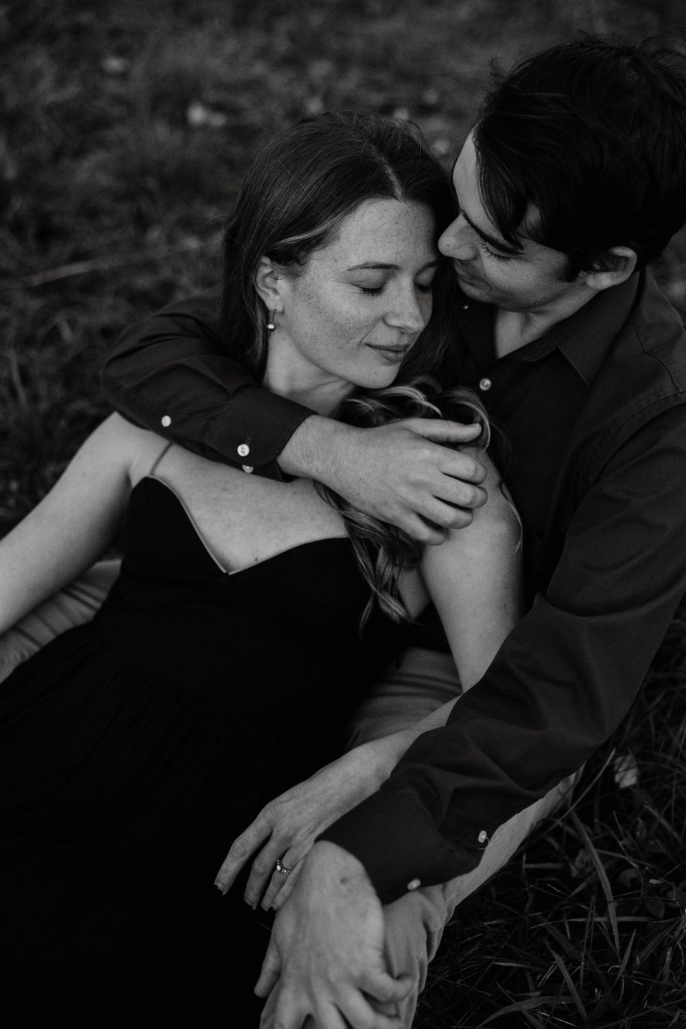 Black and white photo of couple embracing tenderly while sitting together on grass.