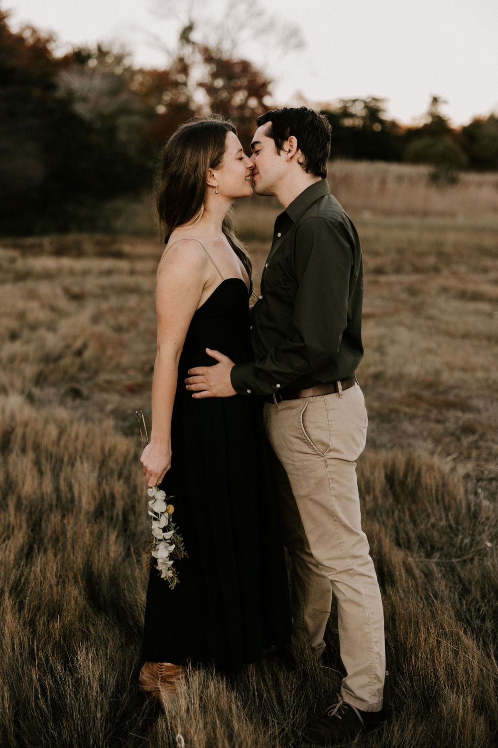 Couple sharing a romantic kiss in a golden field at sunset, woman in black dress holding white flowers.