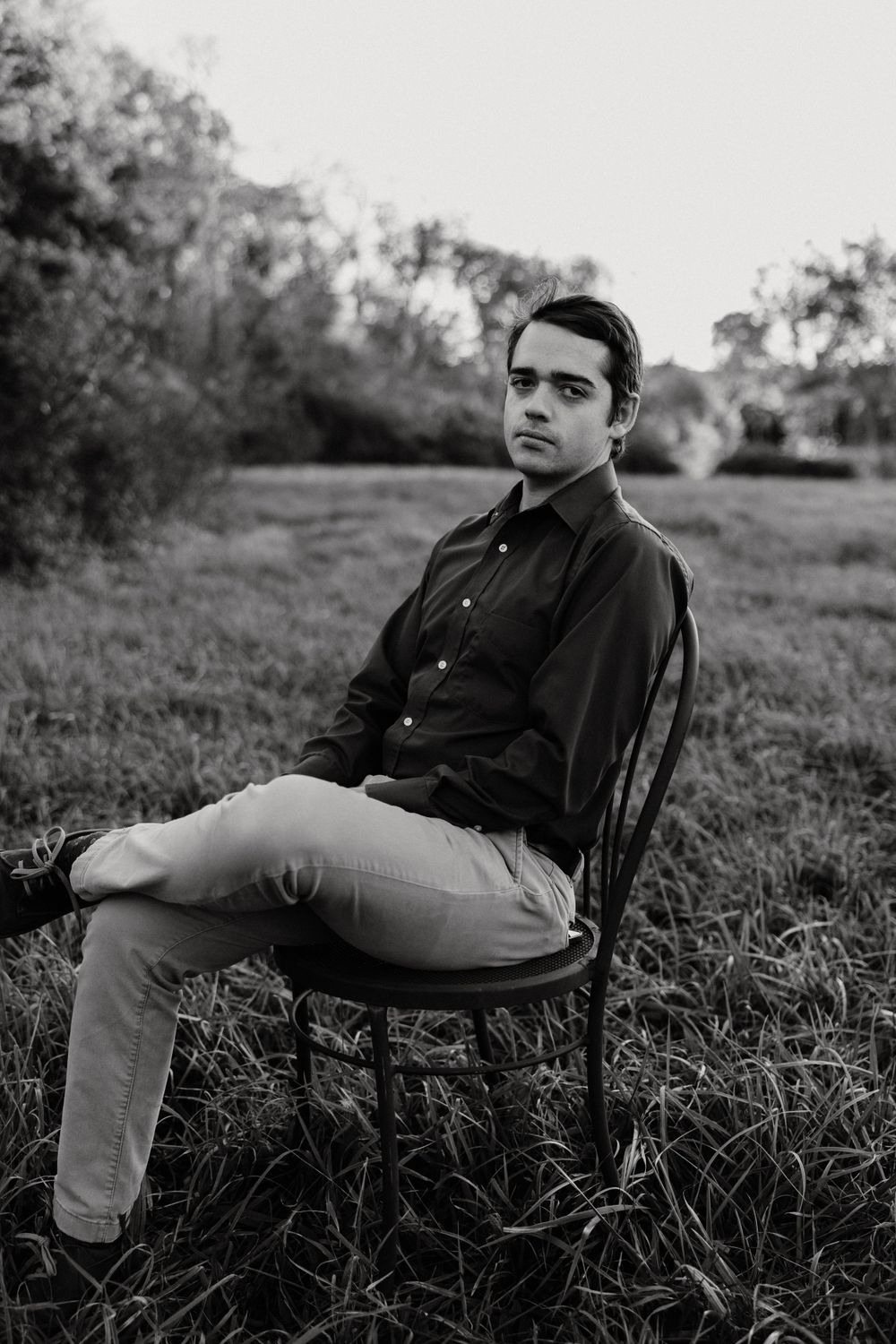 Black and white portrait of a young man sitting on a chair in a grassy field, wearing a dark shirt and light pants. Miranda Photo & Co