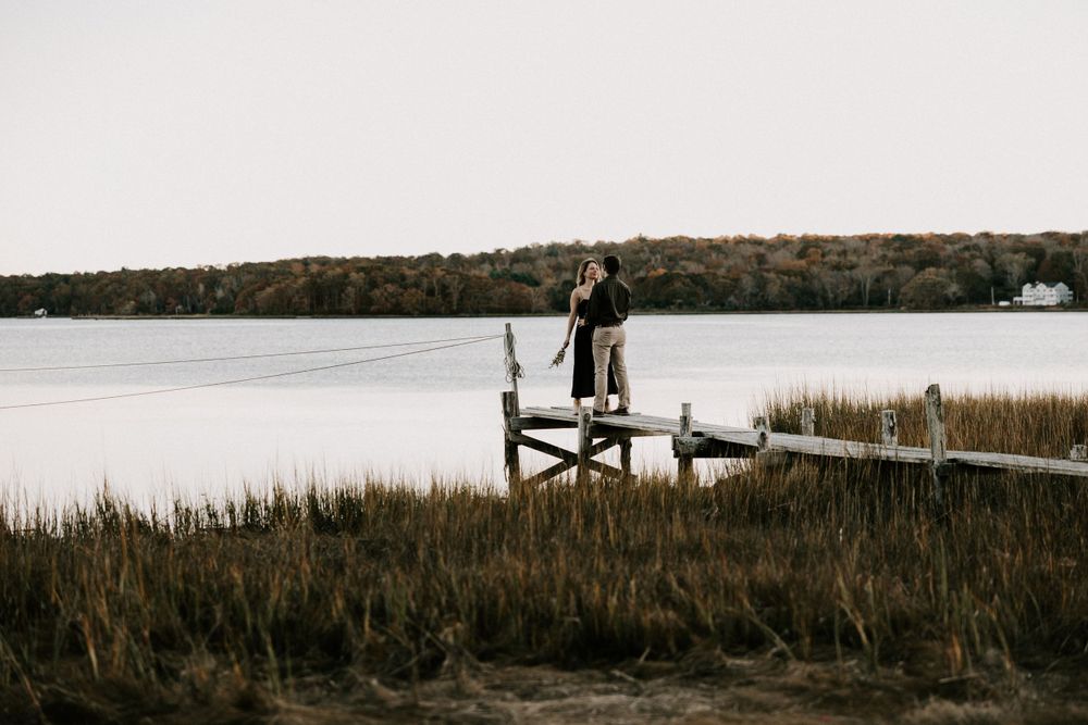 Couple standing on a weathered wooden dock overlooking a calm lake surrounded by marsh grass.