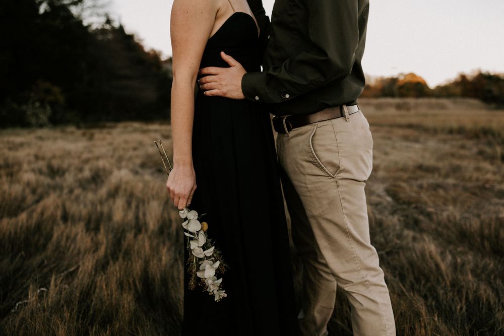 Couple embracing in golden field at sunset, woman holding white eucalyptus bouquet.