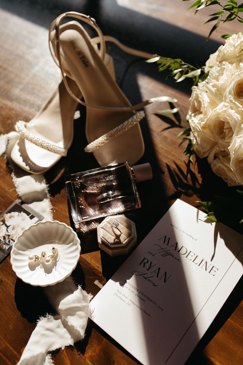 Wedding details with champagne colored heels, white roses, and invitation card on wooden table.