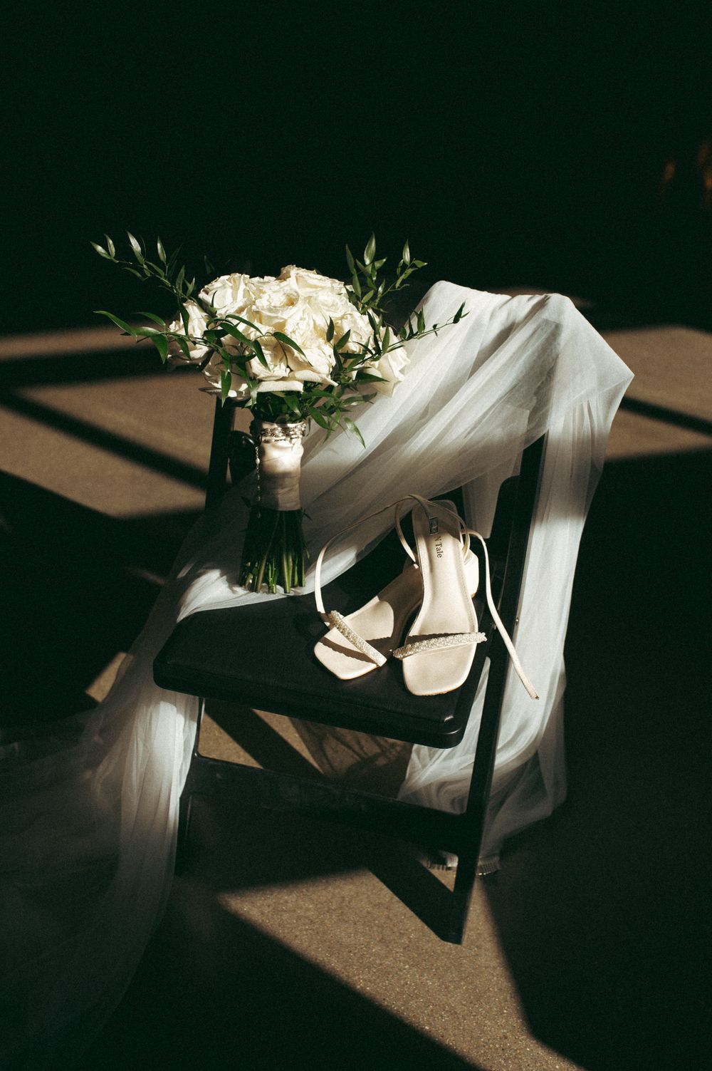 Wedding bouquet and bridal shoes arranged on dark chair with dramatic shadows.