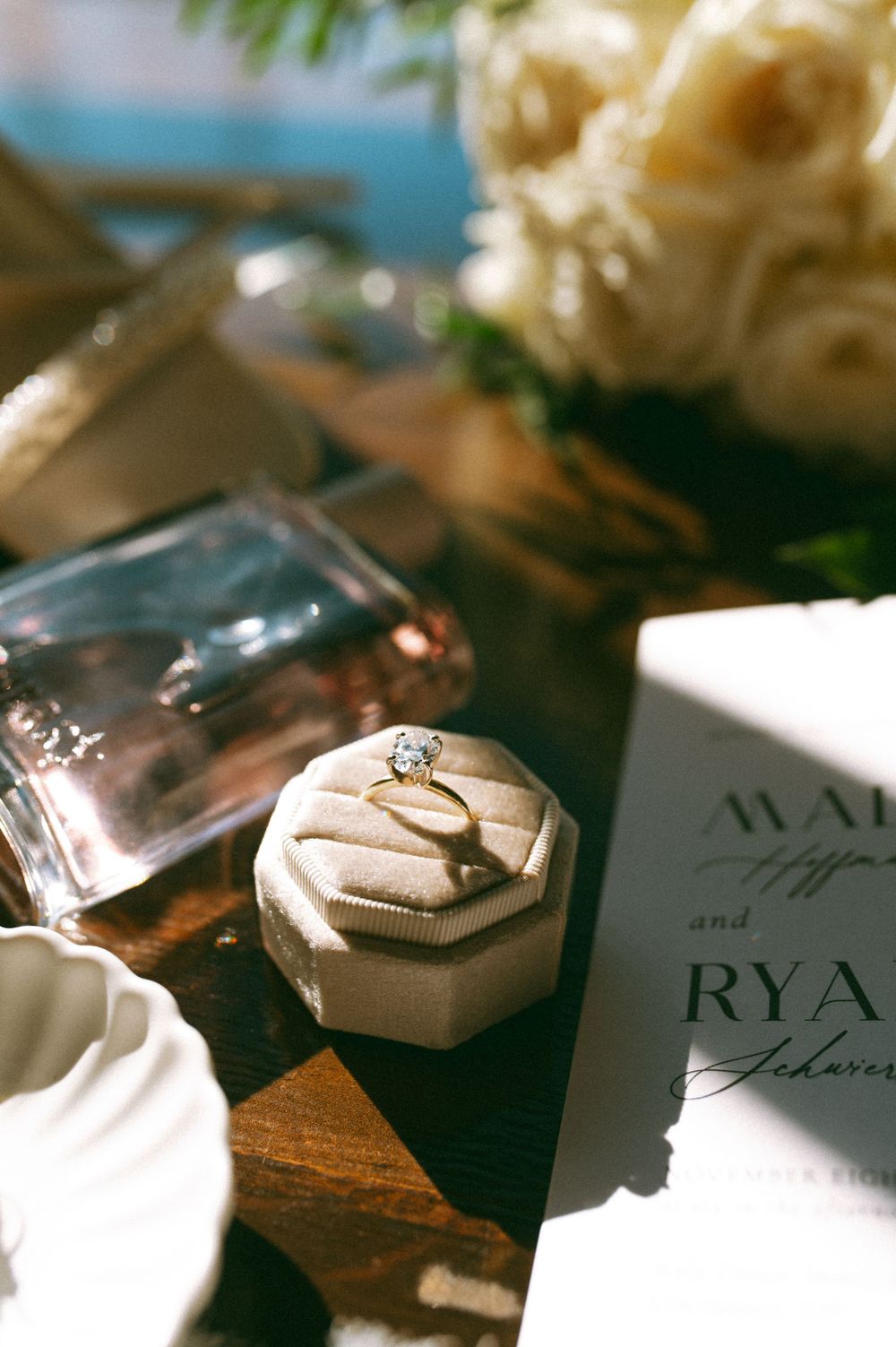 Wedding details showing an engagement ring in a hexagonal box next to white flowers and invitation card.