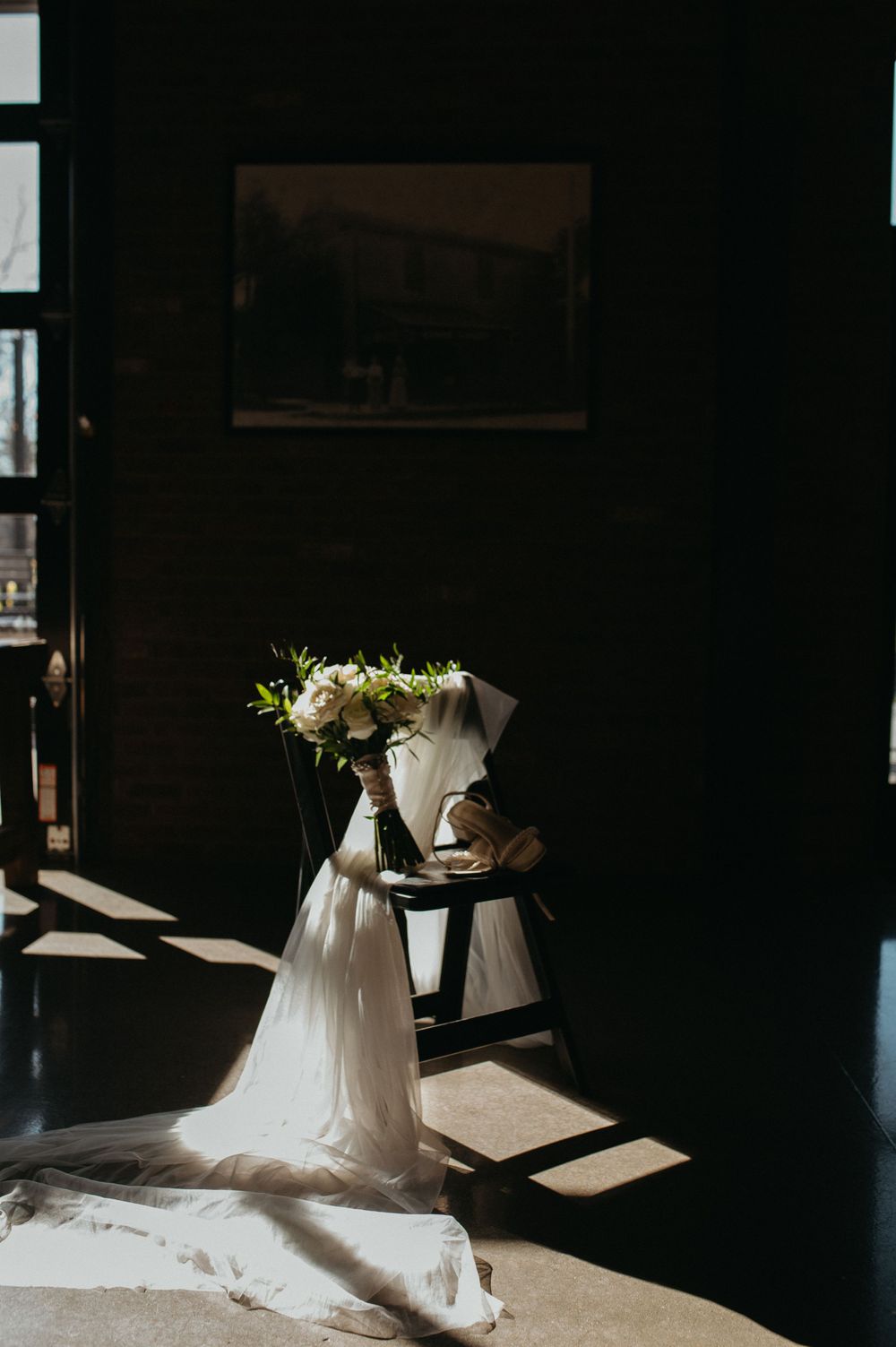 White wedding dress and bouquet artistically draped over chair in dramatic lighting.