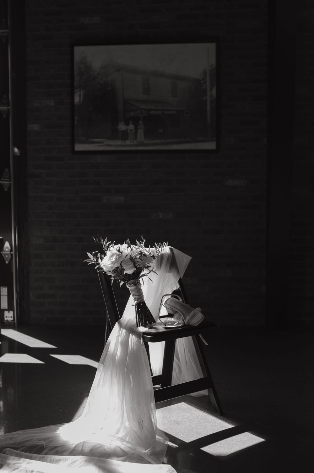 Black and white photo of wedding dress and bouquet on chair with framed artwork in background.