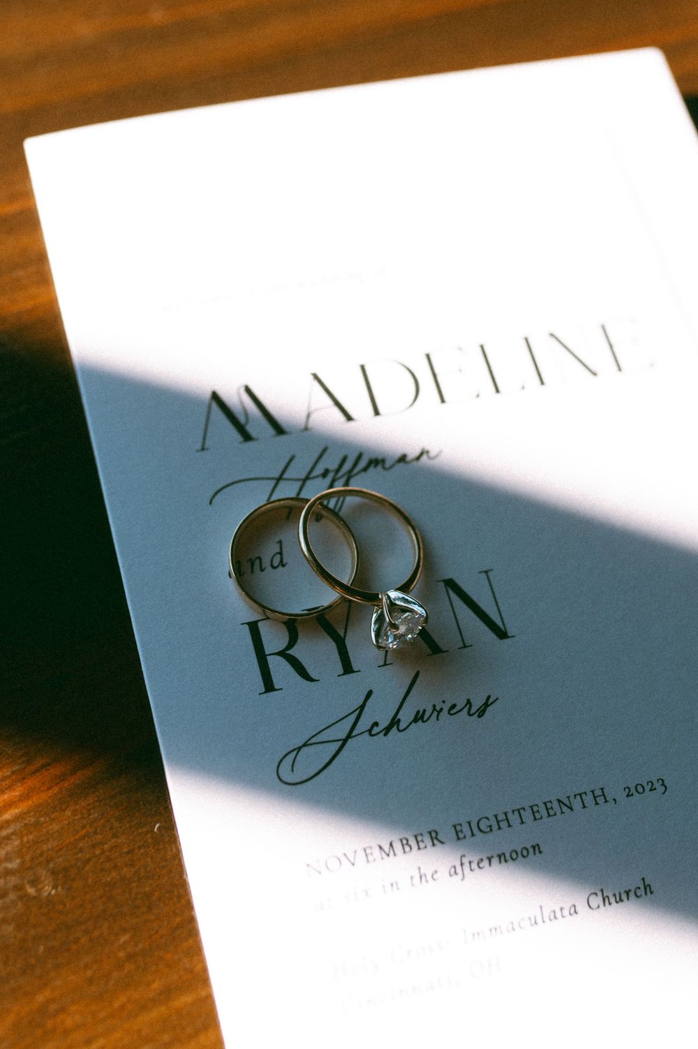 Wedding invitation with engagement rings casting dramatic shadows on wooden surface.