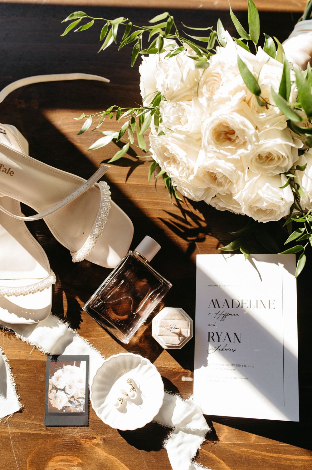Wedding details flatlay with white roses, invitation, shoes and perfume on wooden surface.