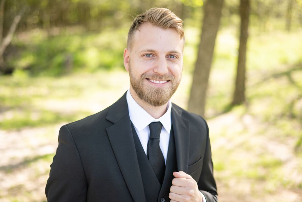 Professional portrait of person in black suit and tie outdoors in wooded setting.
