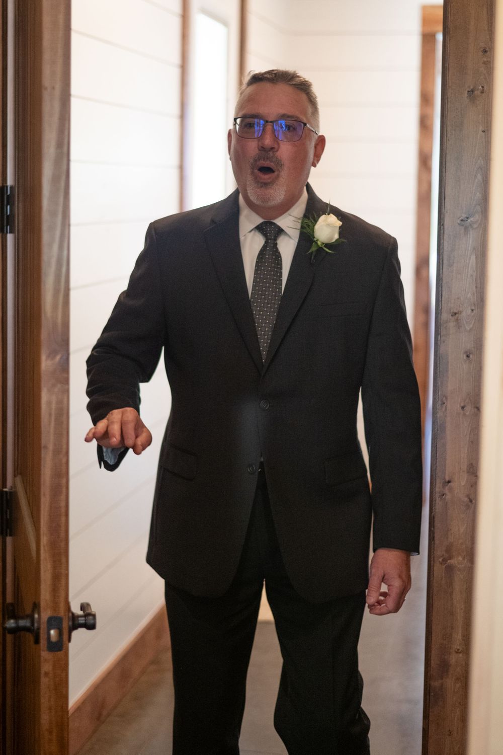 Person in formal attire expressing surprise in hallway wearing glasses and boutonniere.
