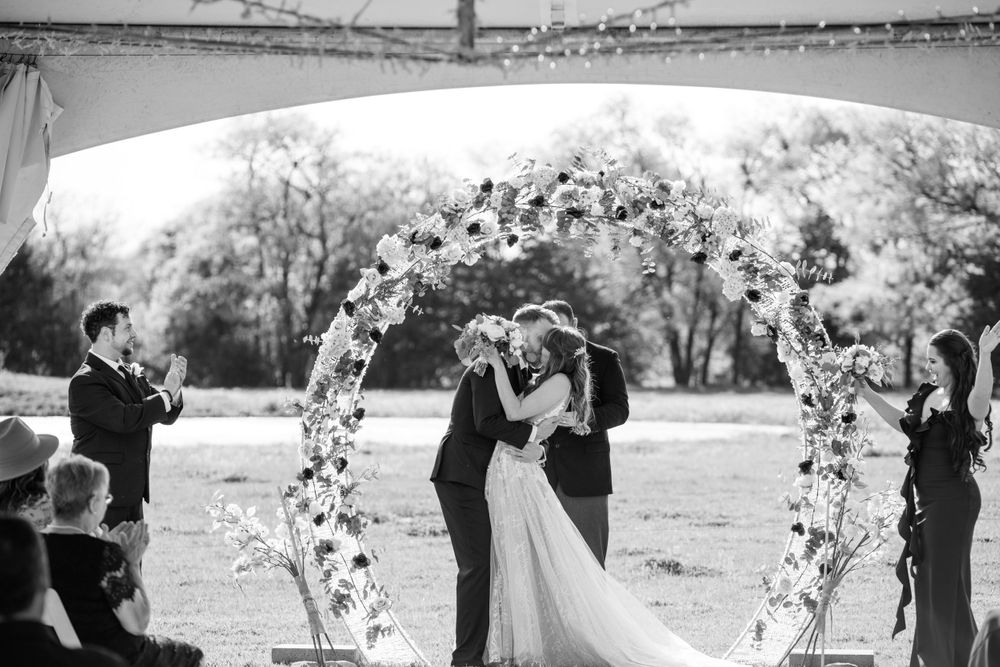 A romantic wedding ceremony under a floral arch with guests watching at an outdoor venue.
