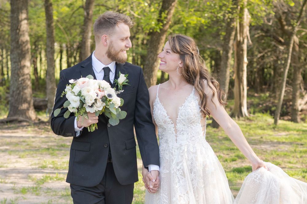 Wedding couple walking together outdoors holding bouquet in wooded setting.