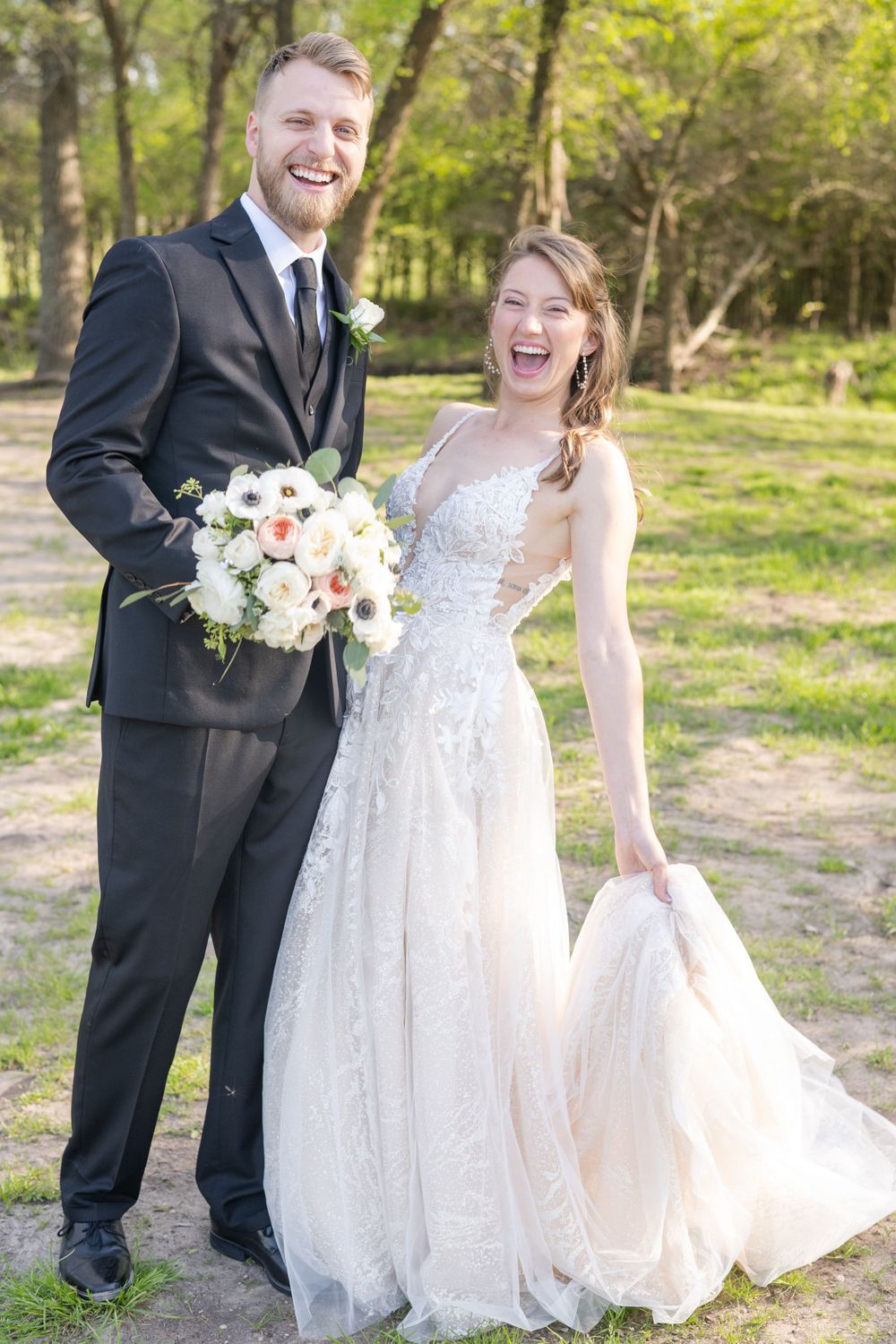 A joyful wedding couple sharing a laugh while holding a bouquet in a wooded setting.