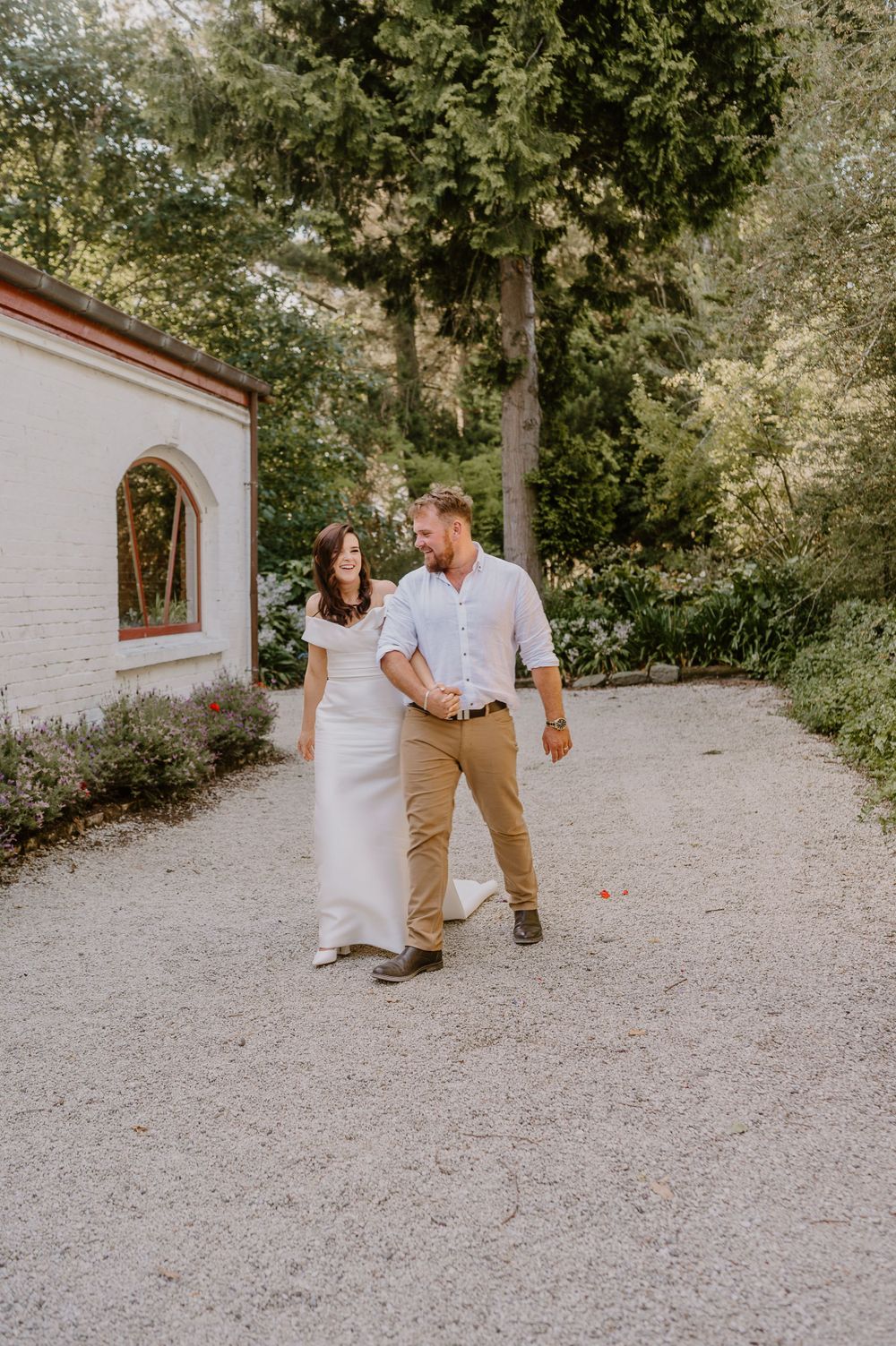 Couple walking together on a gravel path by a white building surrounded by greenery.
