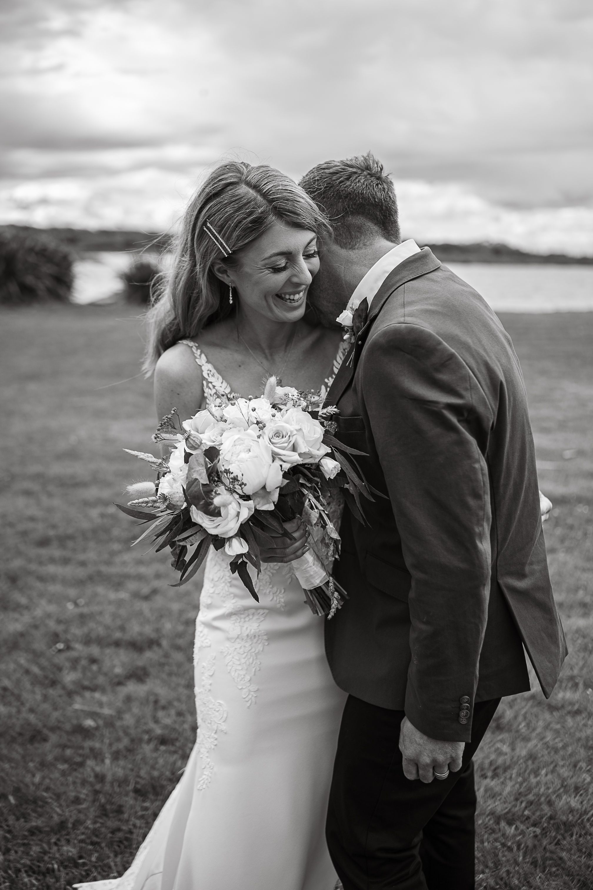 Black and white wedding portrait with couple embracing on coastal cliff.