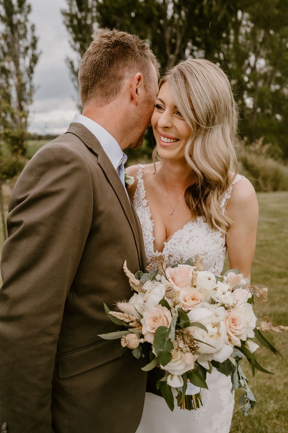 Wedding couple sharing tender moment with blush rose bouquet against pine trees.