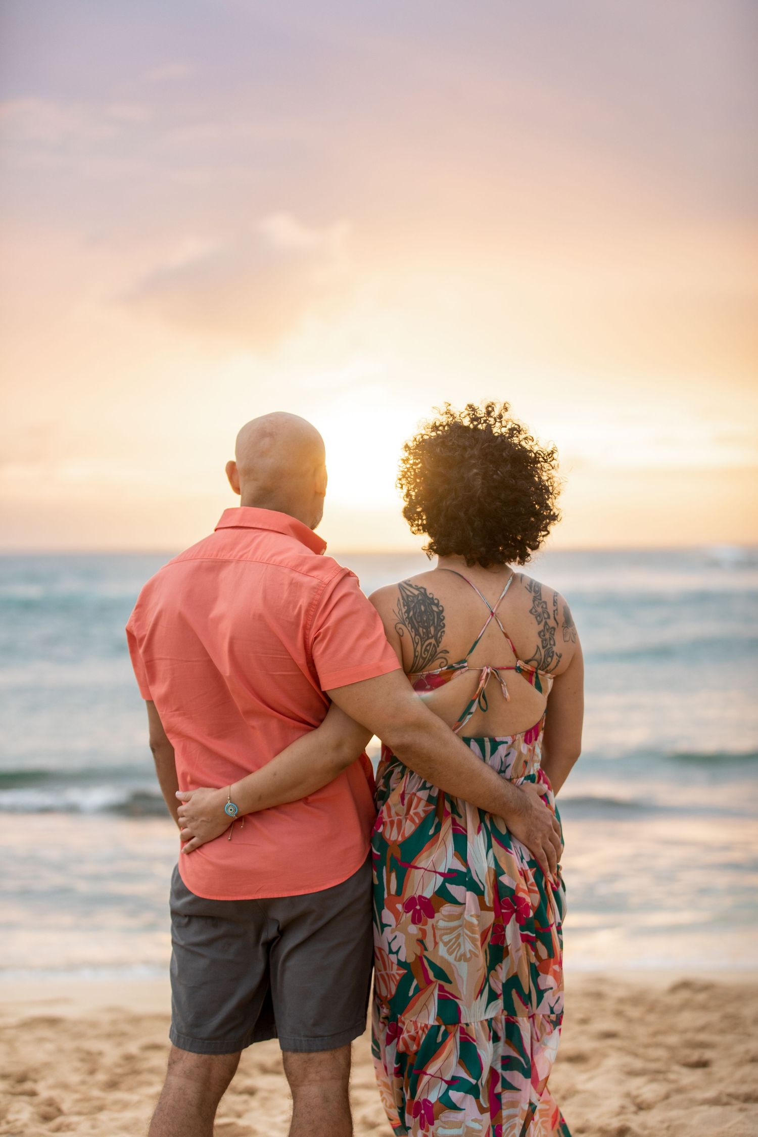 A couple embraces while watching sunset on a beach in tropical attire.