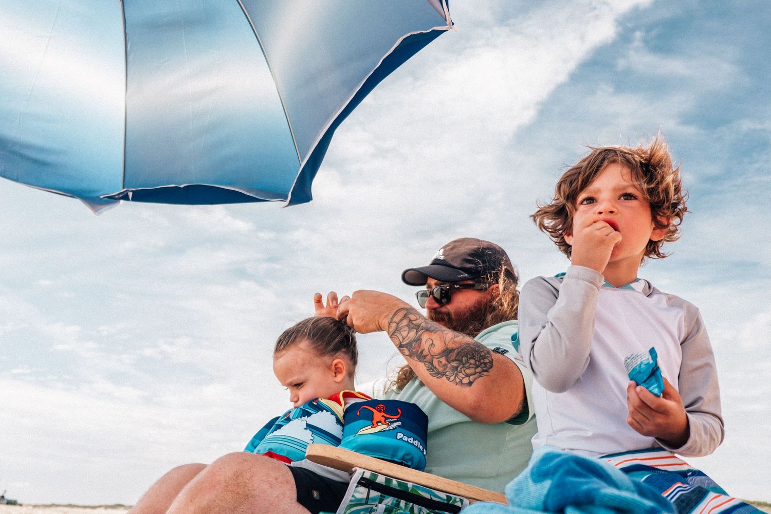 dad putting son's hair into a bun and boy eating chips at sandbridge beach in virginia