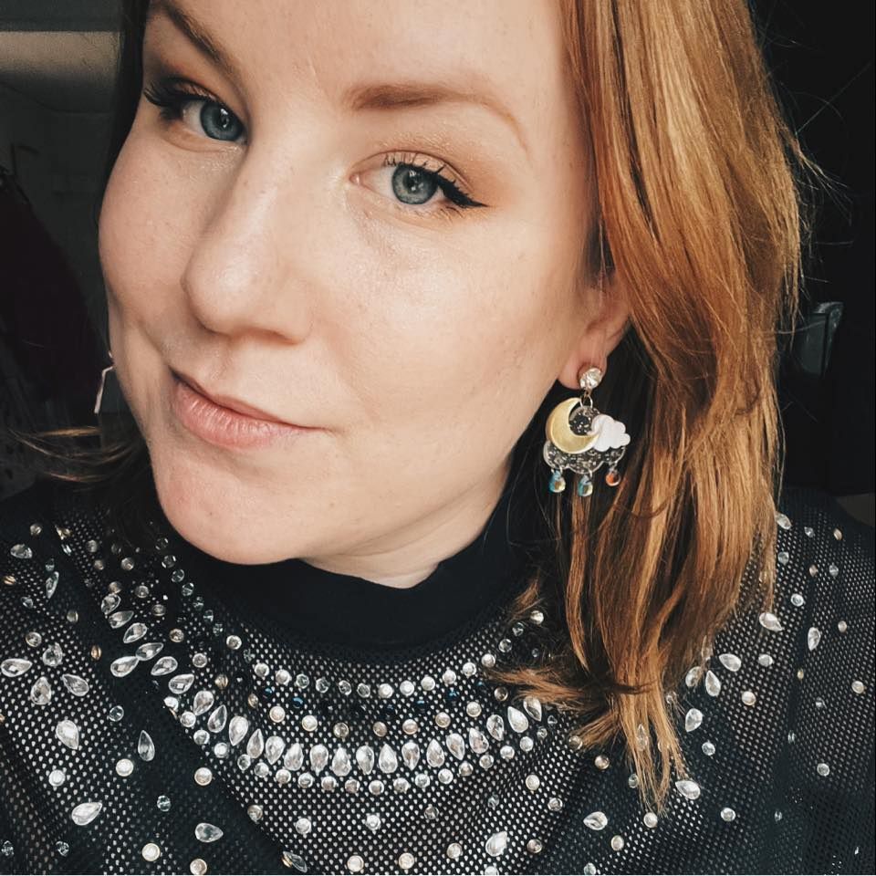Woman with red hair wearing ornate chandelier earrings and a black beaded neckline top, taking a selfie.