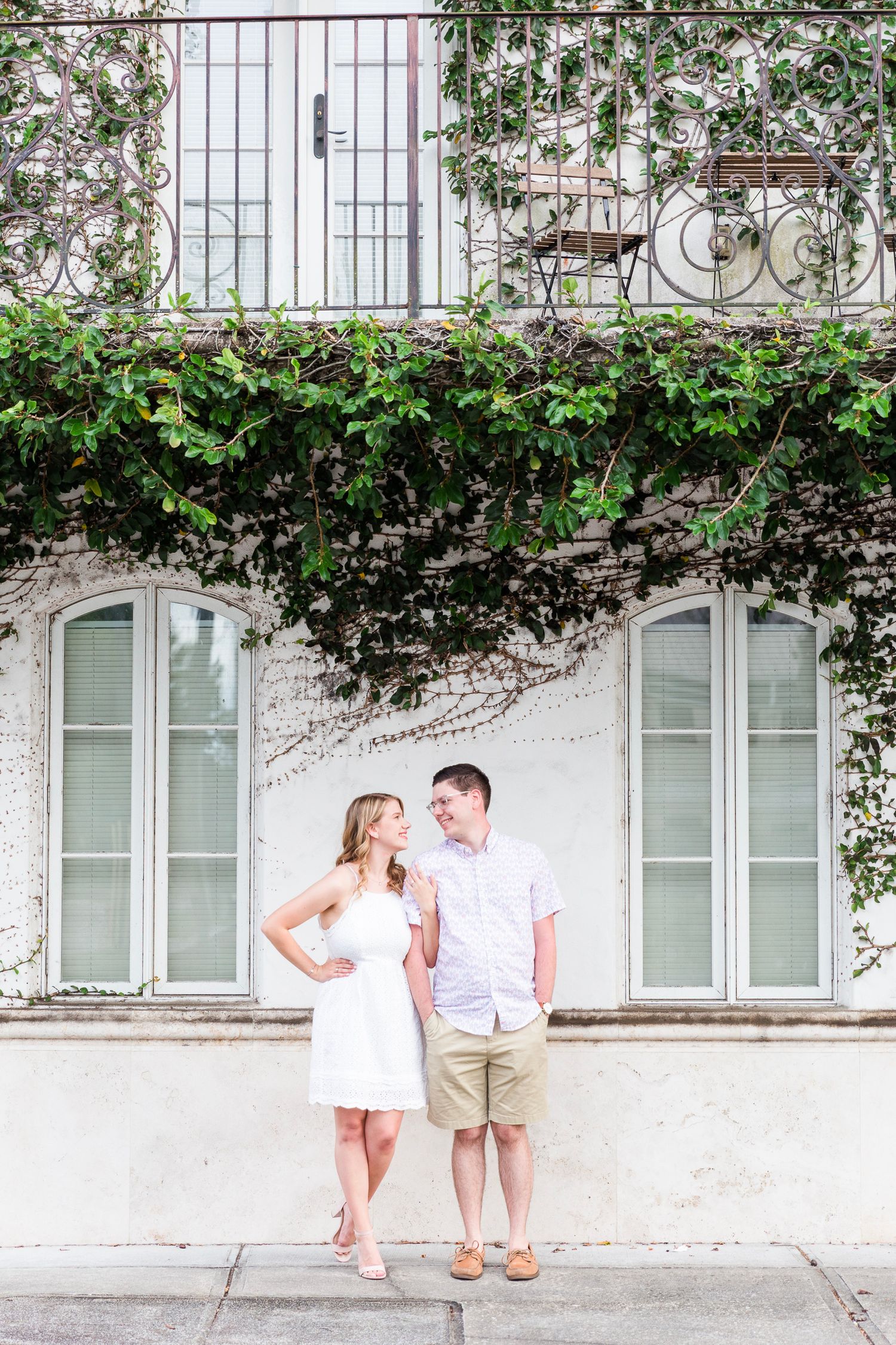 Romantic couple poses against white wall with climbing vines and iron railings.