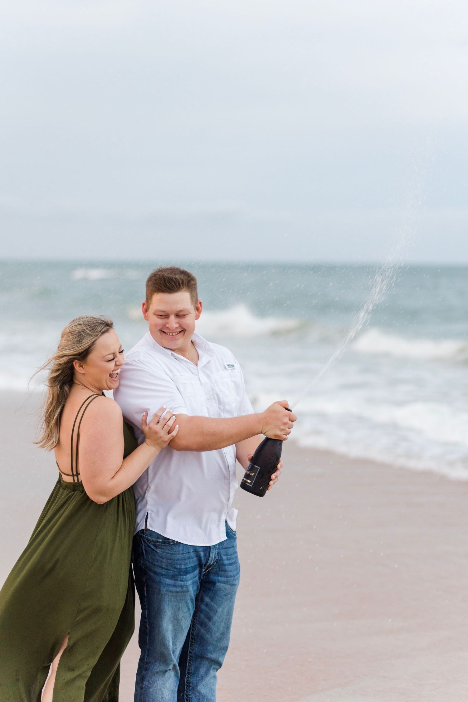 Playful couple popping champagne on sandy beach during sunset celebration.