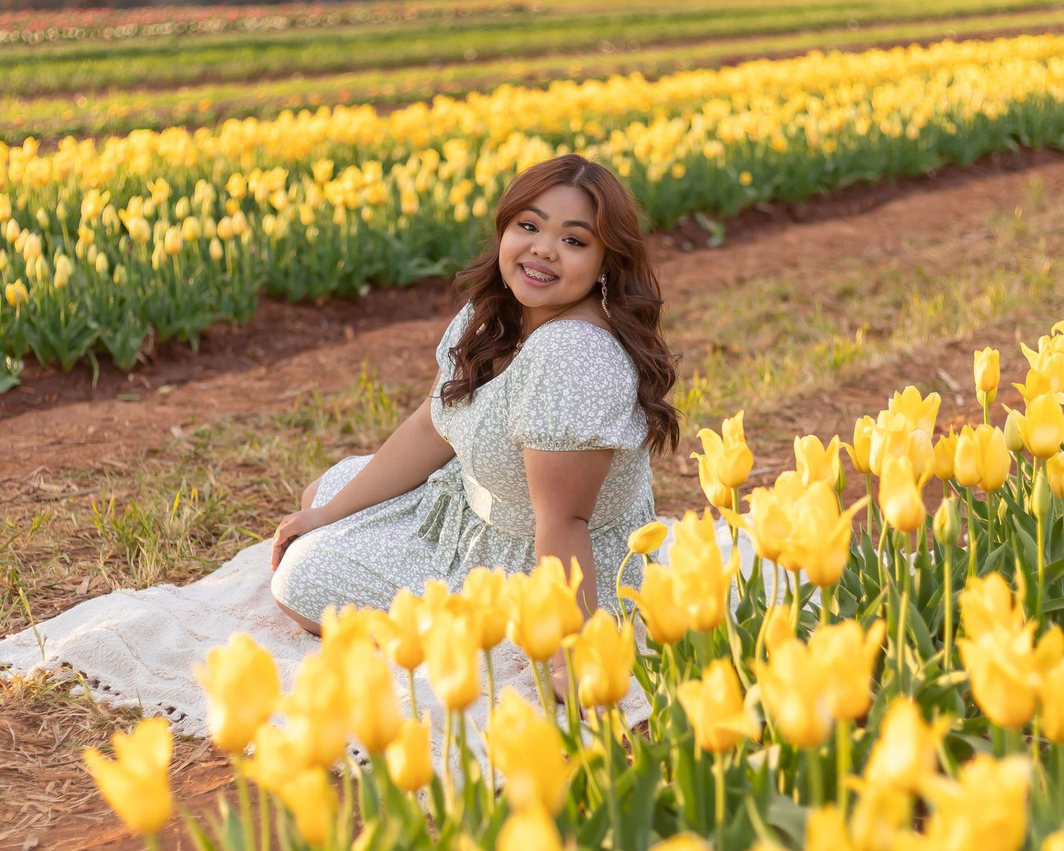 Person in a white dress sits among rows of vibrant yellow tulip flowers during golden hour in a garden field.