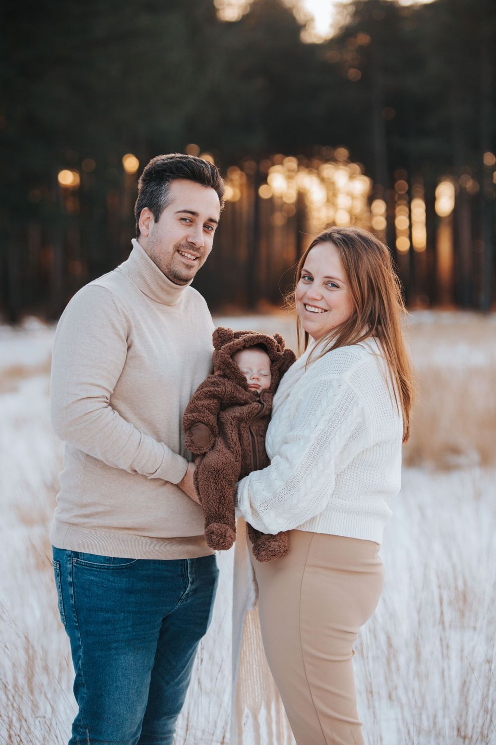 Couple smiling while holding brown teddy bear in snowy field with forest background.