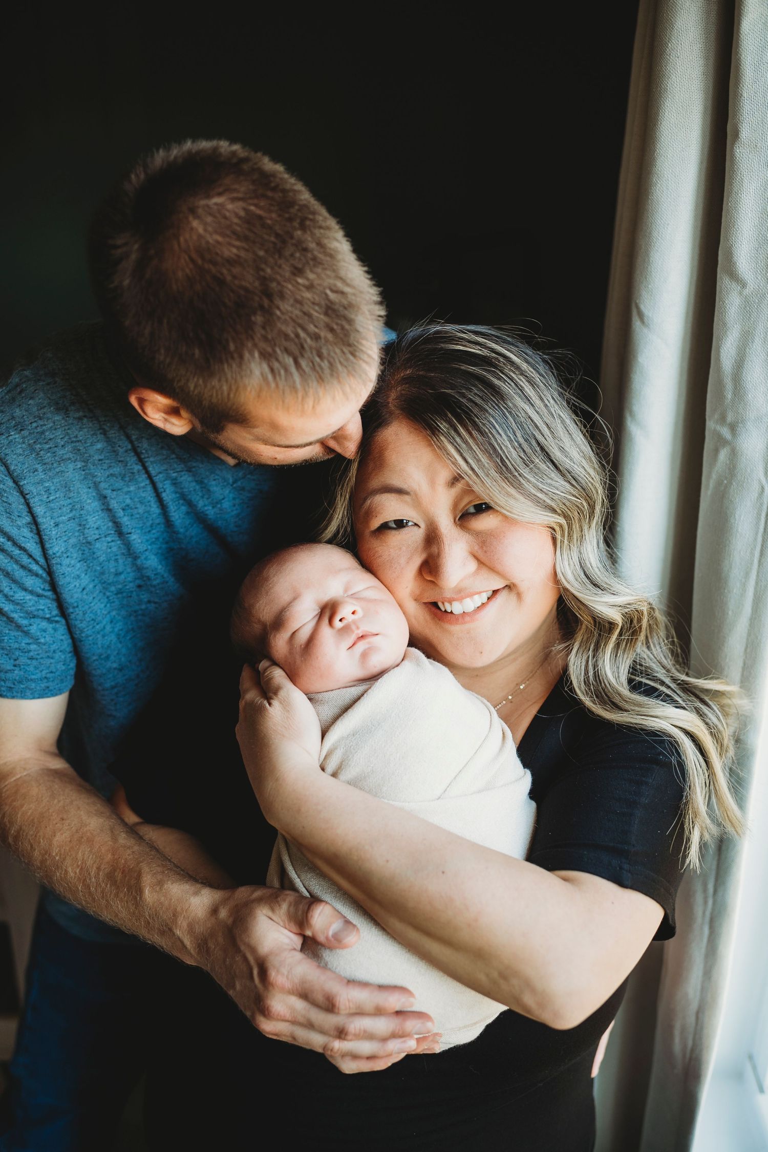 Smiling new parents holding their sleeping swaddled newborn baby near a bright window.