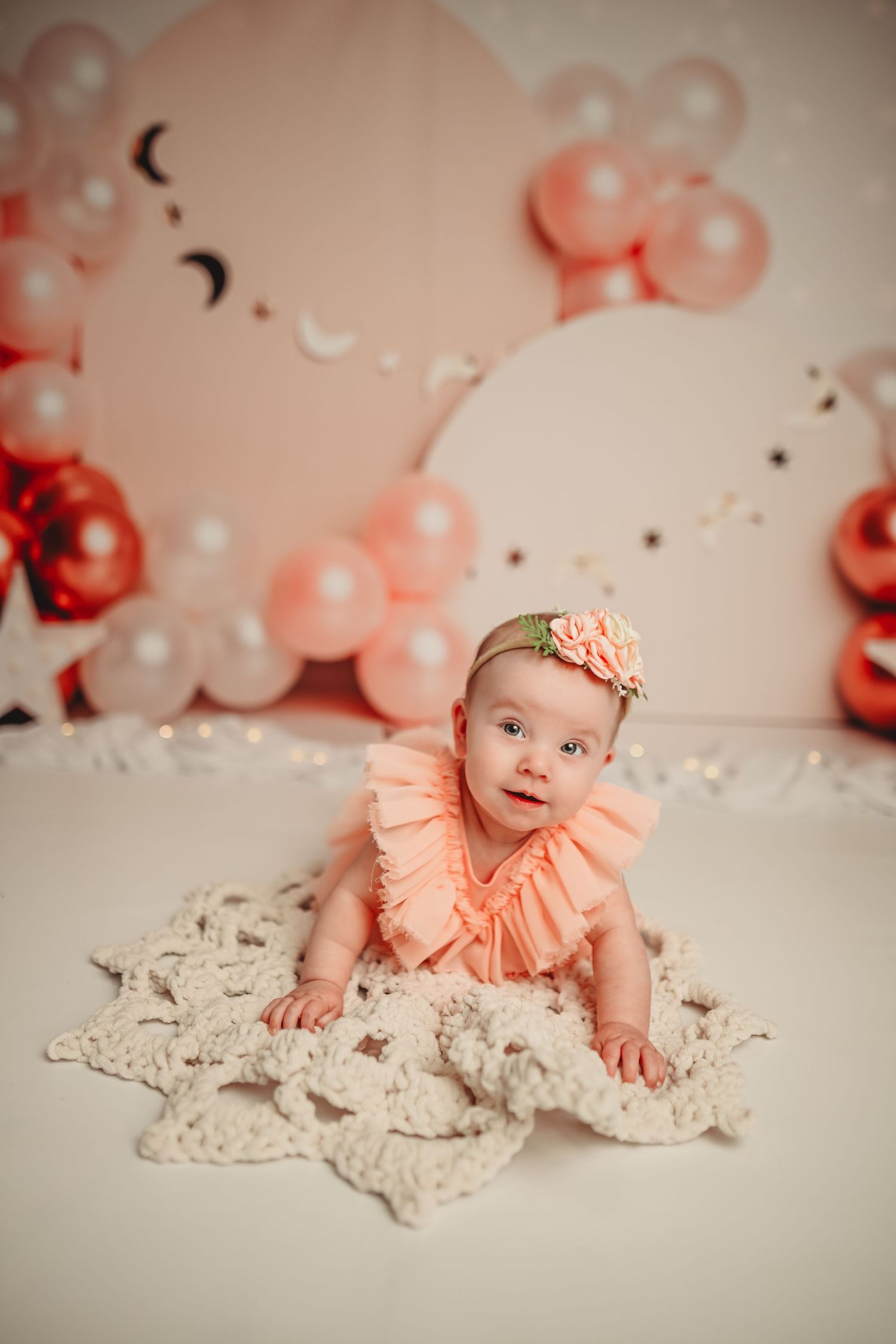 Baby in peach dress lying on lace blanket with pink balloon decorations in background.