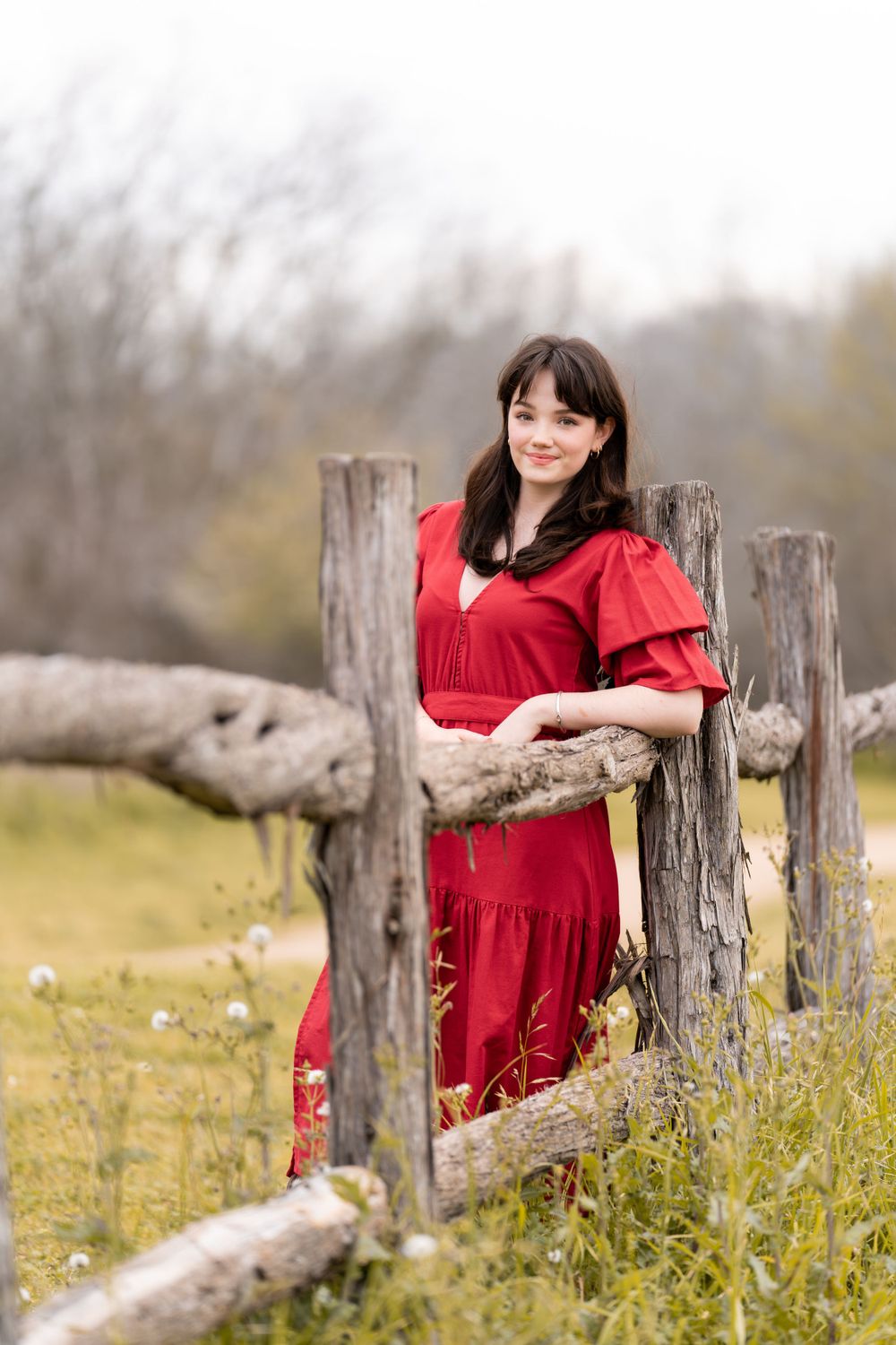 Person leaning against weathered fence posts wearing a flowing red dress in a pastoral field.