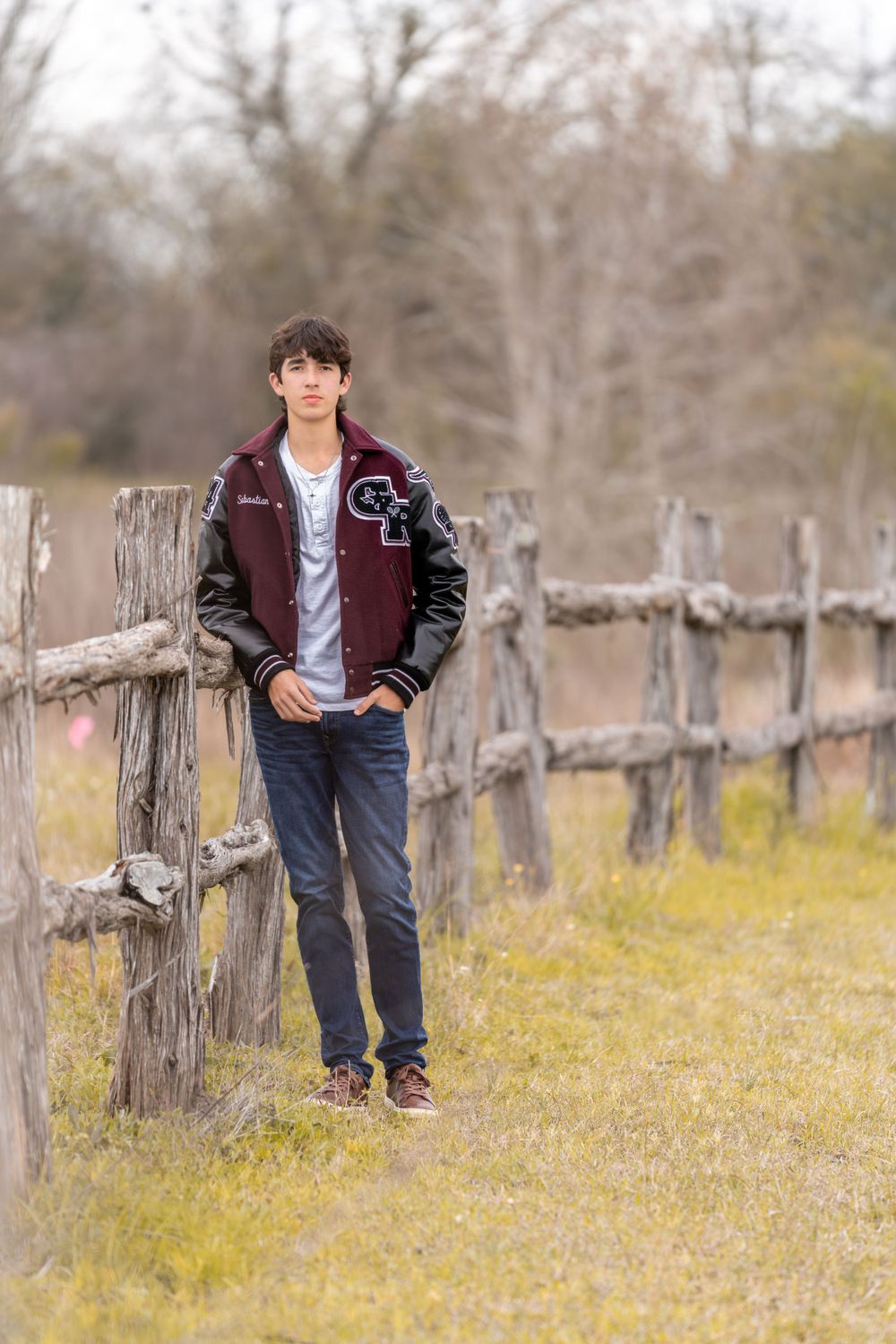 Student in varsity jacket leaning against rustic wooden fence in countryside setting.