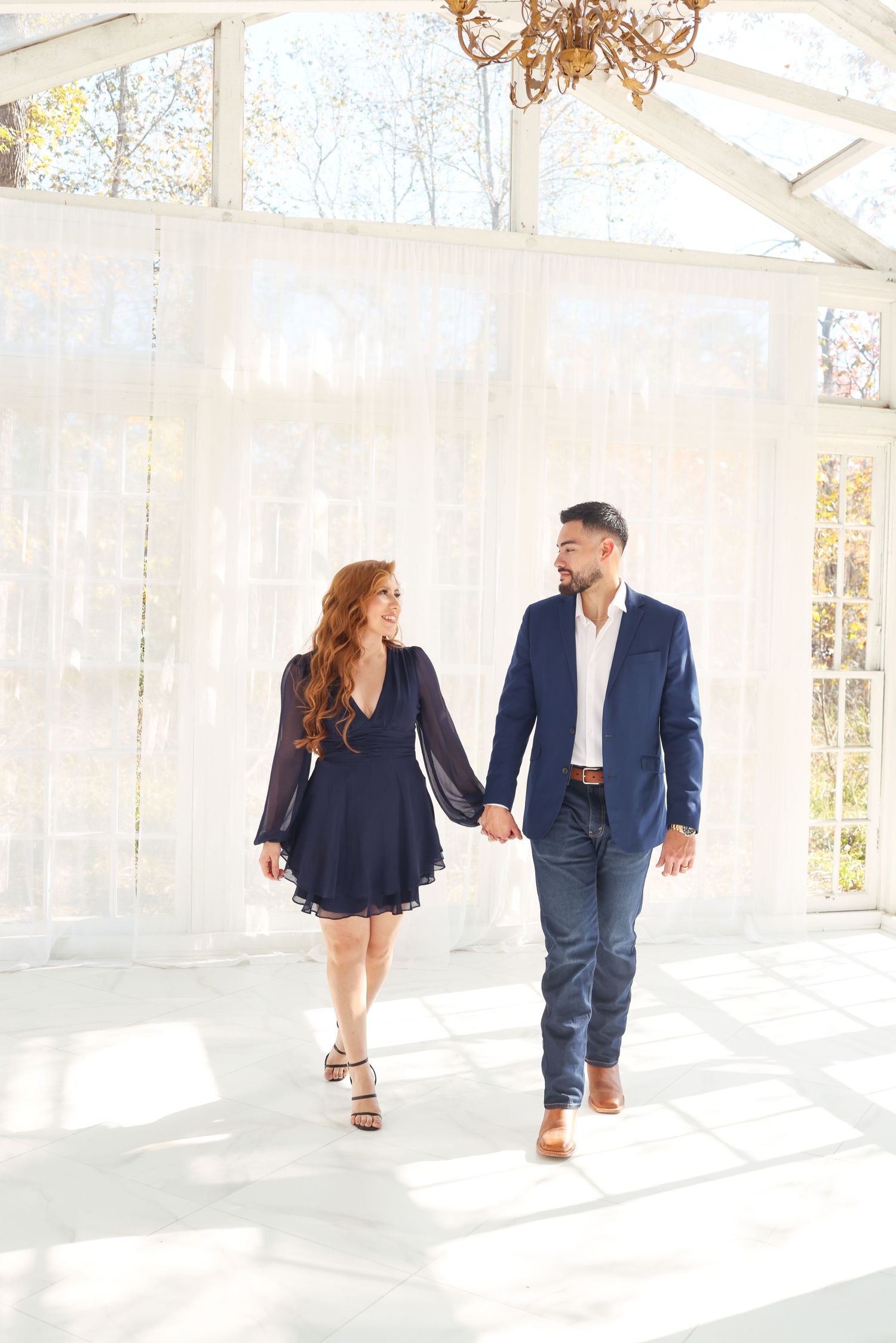 Couple walking hand in hand through sunlit white conservatory space.