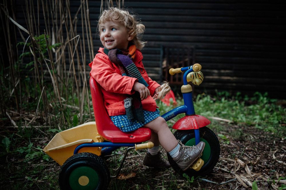 A child in a red coat rides a yellow toy tractor in a garden setting.