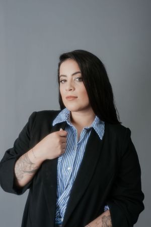 Professional headshot featuring striped button-up shirt and black blazer with visible tattoo details.